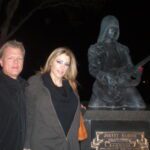 Blond man with a glammed up blond woman posing near a statue of a person playing a guitar