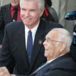 Bespectacled elder man posing with a white-haired man in a formal suit